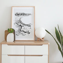 Load image into Gallery viewer, A black and white illustrated print of Kaikōura laid out on white paper within a light wooden frame hanging leaning against a white wall on top of a modern wooden and white console.